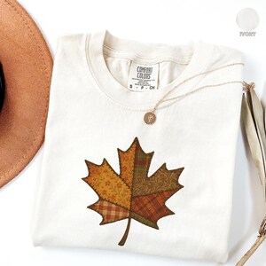 Op de afbeelding: Ivoor kleurig T-shirt met een groot patchwork esdoornblad ontwerp in tinten oranje, geel en groen. Een gouden ketting met hanger ligt op het shirt. Het label luidt "Comfort Colors".