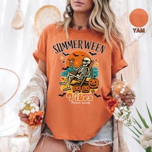 May include: Orange t-shirt with the text "Summerween Vibes Forever Spooky". The graphic features a skeleton relaxing on a beach chair with pumpkins and bats. The shirt is a yam color.