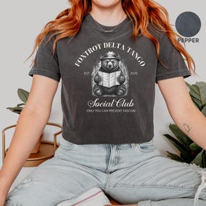 Puede incluir: Una camiseta gris oscuro recortada con un gr&aacute;fico blanco de un oso con un sombrero leyendo un mapa. El texto "Fox Trot Delta Tango Est 2025" est&aacute; por encima del oso y el texto "Social Club Only You Can Prevent Fascism" est&aacute; por debajo del oso.