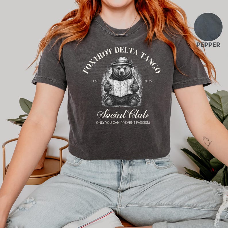 Puede incluir: Una camiseta gris oscuro recortada con un gr&aacute;fico blanco de un oso con un sombrero leyendo un mapa. El texto "Fox Trot Delta Tango Est 2025" est&aacute; por encima del oso y el texto "Social Club Only You Can Prevent Fascism" est&aacute; por debajo del oso.