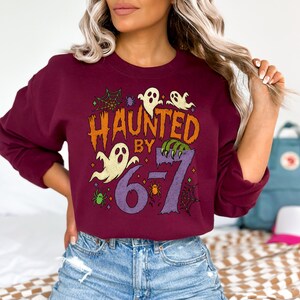 May include: A maroon sweatshirt with the text "HAUNTED BY 67" in orange and purple. The design includes ghost illustrations, spiderwebs, and spiders, perfect for Halloween. The sweatshirt is a crew neck style.