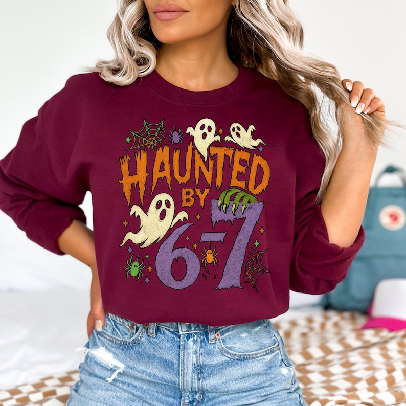 May include: A maroon sweatshirt with the text "HAUNTED BY 67" in orange and purple. The design includes ghost illustrations, spiderwebs, and spiders, perfect for Halloween. The sweatshirt is a crew neck style.