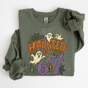 May include: Olive green sweatshirt with a Halloween-themed graphic. The design features the text "HAUNTED BY 67" in orange and purple, along with ghost illustrations, spiderwebs, and spiders. The sweatshirt has long sleeves and a crew neck.