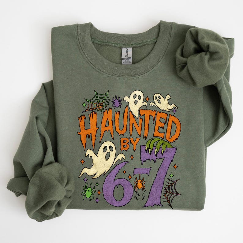 May include: Olive green sweatshirt with a Halloween-themed graphic. The design features the text "HAUNTED BY 67" in orange and purple, along with ghost illustrations, spiderwebs, and spiders. The sweatshirt has long sleeves and a crew neck.