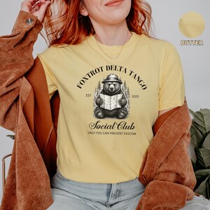 Puede incluir: Una camiseta amarilla con una ilustraci&oacute;n en blanco y negro de un oso con un sombrero y sosteniendo un mapa. El texto "Fox Trot Delta Tango Est 2025 Social Club Only You Can Prevent Fascism" est&aacute; impreso en la camiseta.