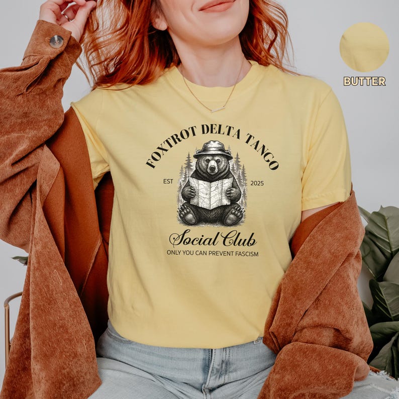 Puede incluir: Una camiseta amarilla con una ilustraci&oacute;n en blanco y negro de un oso con un sombrero y sosteniendo un mapa. El texto "Fox Trot Delta Tango Est 2025 Social Club Only You Can Prevent Fascism" est&aacute; impreso en la camiseta.