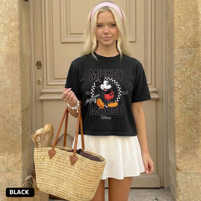 May include: Black t-shirt featuring a Mickey Mouse graphic. The design includes the text "Mickey Mouse" and the numbers "19" and "28". The person is wearing a white skirt and carrying a woven tote bag with brown leather handles.