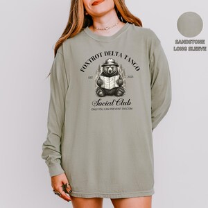 Puede incluir: Una camiseta de manga larga de color arenisca con un gr&aacute;fico de un oso leyendo un libro. El gr&aacute;fico incluye el texto "FOXTROT DELTA TANGO EST 2025 Social Club ONLY YOU CAN PREVENT FASCISM".