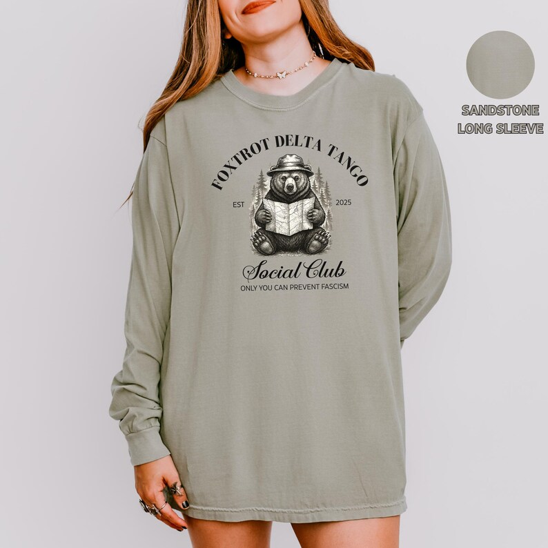 Puede incluir: Una camiseta de manga larga de color arenisca con un gr&aacute;fico de un oso leyendo un libro. El gr&aacute;fico incluye el texto "FOXTROT DELTA TANGO EST 2025 Social Club ONLY YOU CAN PREVENT FASCISM".