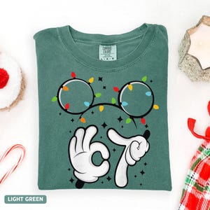 May include: Light green t-shirt with a graphic of Mickey Mouse ears decorated with Christmas lights. The number 67 is displayed with white gloved hands. The words "LIGHT GREEN" are at the bottom.