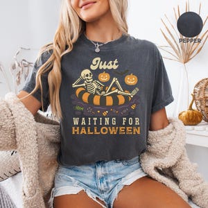 May include: Dark gray t-shirt with the text "Just Waiting for Halloween." The graphic features a skeleton relaxing on an orange and black inner tube with pumpkins and a bat. The shirt is a relaxed fit.