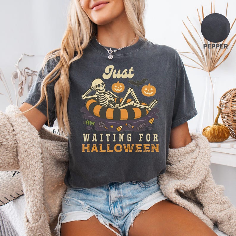 May include: Dark gray t-shirt with the text "Just Waiting for Halloween." The graphic features a skeleton relaxing on an orange and black inner tube with pumpkins and a bat. The shirt is a relaxed fit.