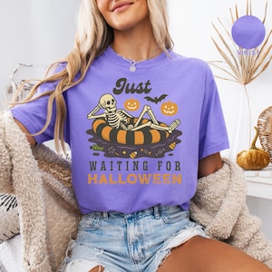 May include: A violet t-shirt with a Halloween-themed graphic. The design features a skeleton relaxing on an orange and black inner tube, with pumpkins and bats. The text reads "Just Waiting for Halloween."