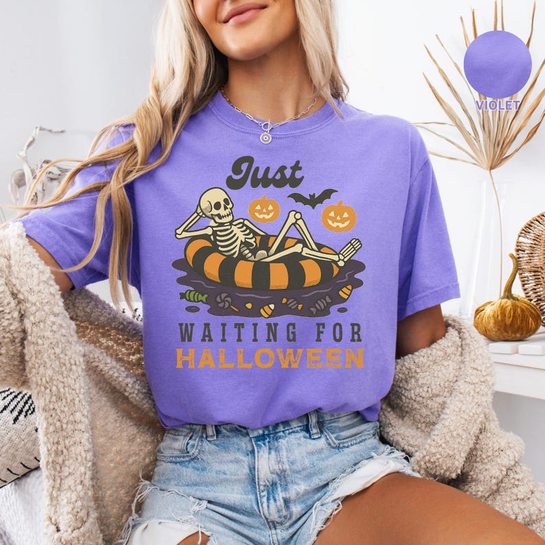 May include: A violet t-shirt with a Halloween-themed graphic. The design features a skeleton relaxing on an orange and black inner tube, with pumpkins and bats. The text reads "Just Waiting for Halloween."