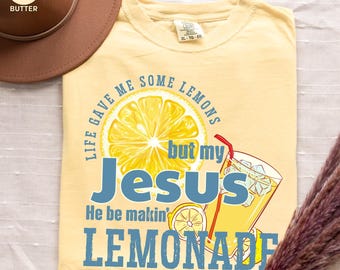 May include: Butter-colored t-shirt with the text "Life gave me some lemons but my Jesus He be makin' lemonade." The design includes a lemon slice and a glass of lemonade. A brown hat is in the upper left corner.