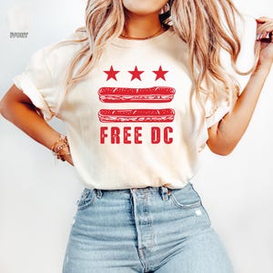 Könnte beinhalten: Elfenbeinfarbenes T-Shirt mit rotem Grafikdesign. Das Design umfasst drei rote Sterne über zwei roten Sandwich-Illustrationen, mit den Worten "FREE DC" darunter in Rot gedruckt. Das Shirt ist ein lässiger Stil.
