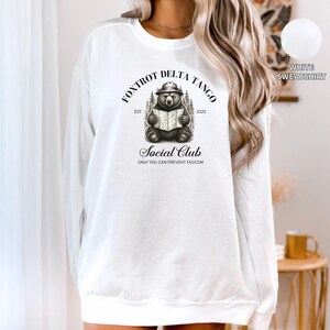 Puede incluir: Sudadera blanca con una ilustraci&oacute;n en blanco y negro de un oso leyendo un libro. El texto "Fox Trot Delta Tango Social Club Est 2025 Only You Can Prevent Fascism" est&aacute; impreso en la sudadera.