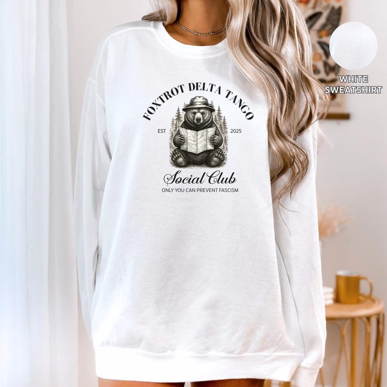 Puede incluir: Sudadera blanca con una ilustraci&oacute;n en blanco y negro de un oso leyendo un libro. El texto "Fox Trot Delta Tango Social Club Est 2025 Only You Can Prevent Fascism" est&aacute; impreso en la sudadera.