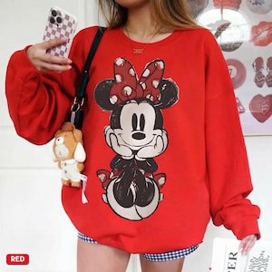 Puede incluir: Una sudadera roja con un dise&ntilde;o de Minnie Mouse, que incluye un lazo de lunares rojos y blancos. La sudadera de manga larga tiene cuello redondo. La palabra "RED" est&aacute; impresa en un rect&aacute;ngulo blanco en la esquina inferior izquierda.