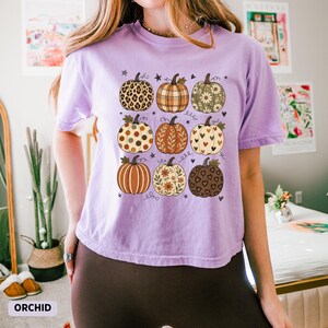 May include: A light purple short-sleeve t-shirt featuring a graphic print of nine pumpkins arranged in a grid. Each pumpkin displays a unique pattern, including leopard print, plaid, floral motifs, polka dots, and hearts, accented by small stars.