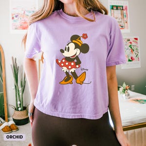 Puede incluir: Camiseta lila con un estampado de Minnie Mouse. El personaje de dibujos animados lleva un vestido rojo de lunares, zapatos amarillos y un sombrero de flores. La palabra "Disney" est&aacute; impresa debajo del gr&aacute;fico. La palabra "ORCHID" est&aacute; impresa en la parte inferior izquierda.
