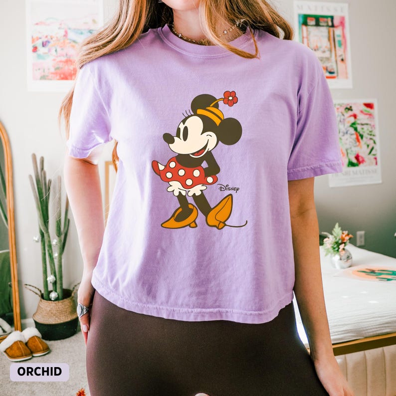 Puede incluir: Camiseta lila con un estampado de Minnie Mouse. El personaje de dibujos animados lleva un vestido rojo de lunares, zapatos amarillos y un sombrero de flores. La palabra "Disney" est&aacute; impresa debajo del gr&aacute;fico. La palabra "ORCHID" est&aacute; impresa en la parte inferior izquierda.