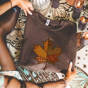 Op de afbeelding: Een bruine sweatshirt met een grote esdoornblad-appliqué. Het blad is gemaakt van verschillende stoffen met patronen in geel, oranje en rood, wat een patchwork-effect geeft. De sweatshirt wordt door handen omhooggehouden.