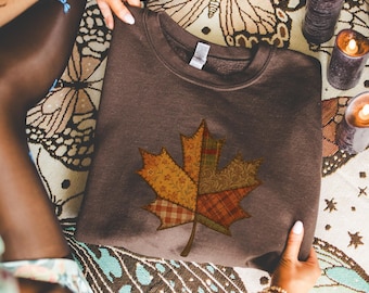 May include: A brown sweatshirt with a large maple leaf appliqué. The leaf is made from various patterned fabrics in yellow, orange, and red, giving a patchwork appearance. The sweatshirt is held up by hands.