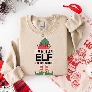 Może przedstawiać: Beżowa bluza z napisem "I'M NOT AN ELF I'M JUST SHORT" w kolorze czarnym, czerwonym, zielonym i żółtym. Projekt zawiera czapkę i stopy elfa. Świąteczny przedmiot tematyczny.