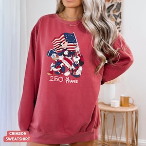 May include: Crimson sweatshirt featuring a patriotic design with cartoon characters holding a flag. The graphic includes the text "250 Years". The sweatshirt is a solid crimson color.
