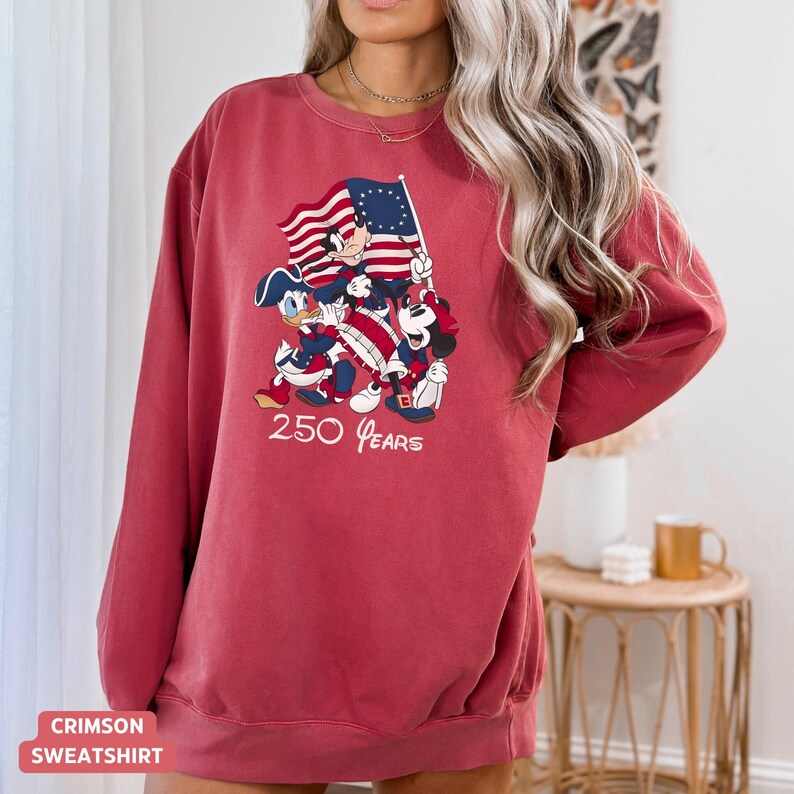 May include: Crimson sweatshirt featuring a patriotic design with cartoon characters holding a flag. The graphic includes the text "250 Years". The sweatshirt is a solid crimson color.