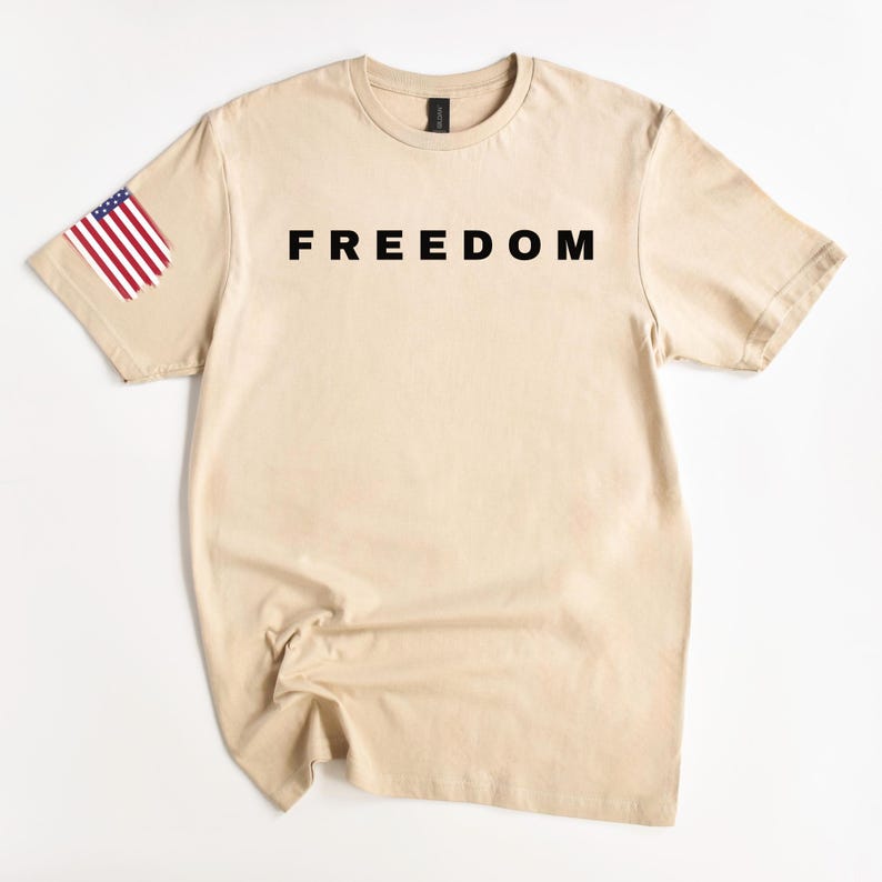 K&ouml;nnte beinhalten: Ein beigefarbenes T-Shirt mit dem Wort "FREEDOM" in Schwarz auf der Brust aufgedruckt. Der linke &Auml;rmel hat ein kleines amerikanisches Flaggen-Design. Das T-Shirt hat einen Rundhalsausschnitt und kurze &Auml;rmel.