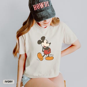 Puede incluir: Camiseta de color marfil con un dibujo de Mickey Mouse. El personaje de dibujos animados lleva pantalones cortos rojos, zapatos amarillos y guantes negros. La camiseta se combina con una gorra negra con la palabra "HOWDY" en rojo.