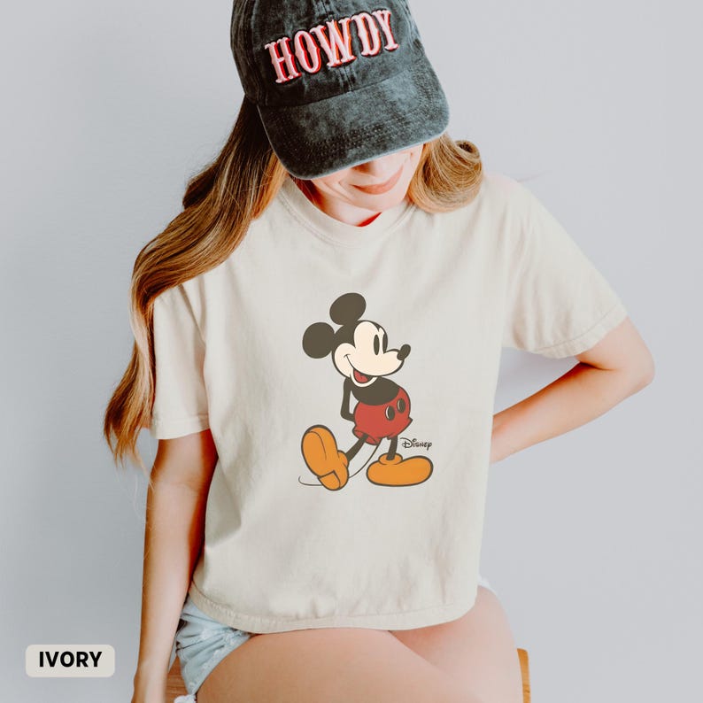 Puede incluir: Camiseta de color marfil con un dibujo de Mickey Mouse. El personaje de dibujos animados lleva pantalones cortos rojos, zapatos amarillos y guantes negros. La camiseta se combina con una gorra negra con la palabra "HOWDY" en rojo.
