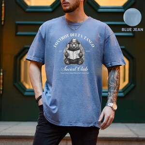 Puede incluir: Una camiseta azul jean con un gr&aacute;fico de un oso con un sombrero leyendo un libro. El texto "Foxtrot Delta Tango Est 2025 Social Club Only You Can Prevent Fascism" est&aacute; impreso en la camiseta.