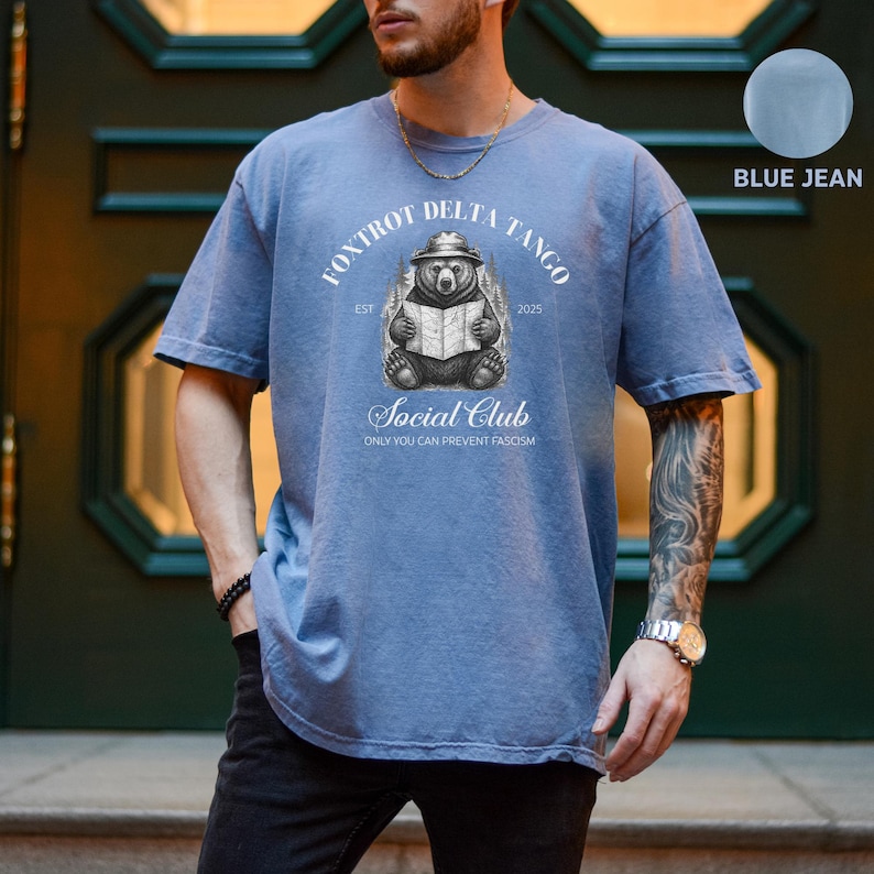 Puede incluir: Una camiseta azul jean con un gr&aacute;fico de un oso con un sombrero leyendo un libro. El texto "Foxtrot Delta Tango Est 2025 Social Club Only You Can Prevent Fascism" est&aacute; impreso en la camiseta.