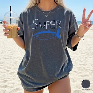 May include: A dark grey t-shirt featuring the word "Super" in white script and a blue fish design. The shirt has a relaxed fit and is labelled "Pepper" in colour.