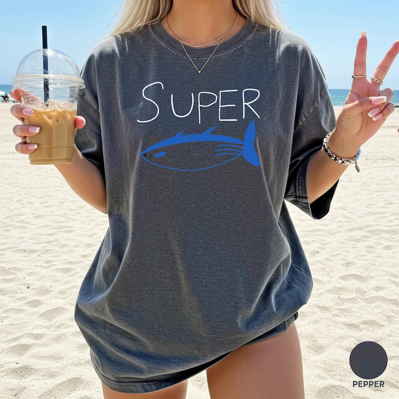 May include: A dark grey t-shirt featuring the word "Super" in white script and a blue fish design. The shirt has a relaxed fit and is labelled "Pepper" in colour.