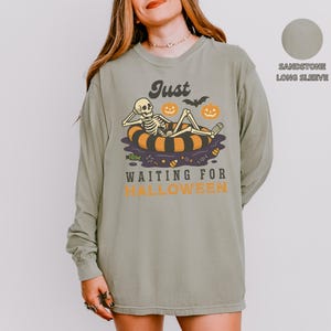 May include: Sandstone long-sleeve shirt with a Halloween graphic. The design features a skeleton relaxing on an inner tube with pumpkins and the text "Just Waiting for Halloween".