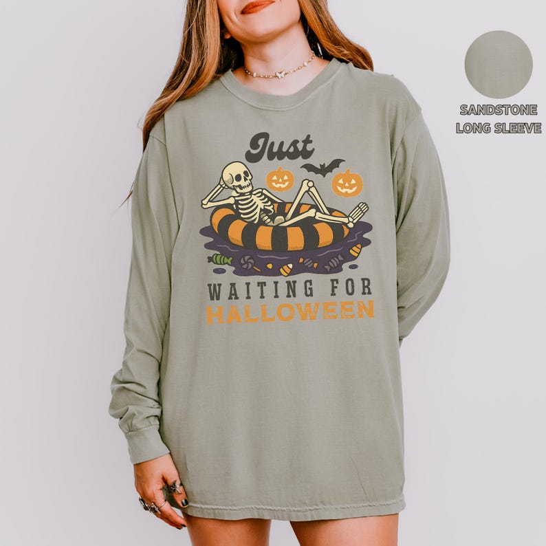 May include: Sandstone long-sleeve shirt with a Halloween graphic. The design features a skeleton relaxing on an inner tube with pumpkins and the text "Just Waiting for Halloween".