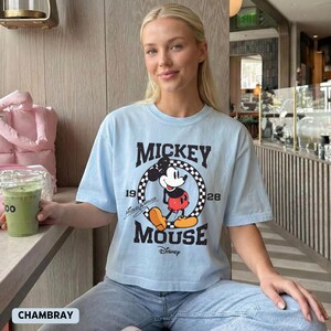 May include: Light blue t-shirt featuring a graphic of Mickey Mouse. The design includes the words "Mickey Mouse" and the years "1928" and "19". The shirt is paired with blue jeans.