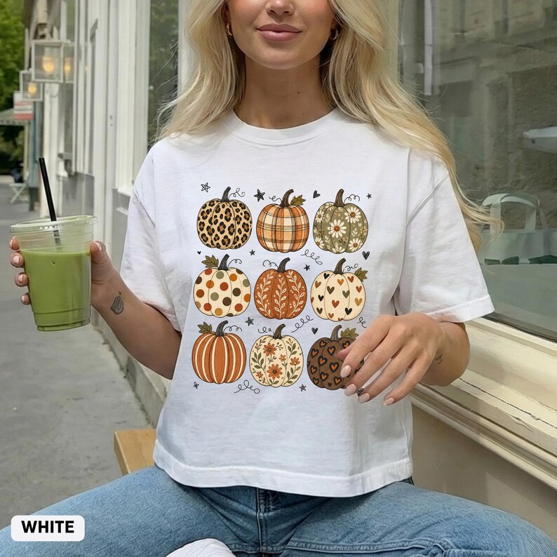 May include: A white short-sleeve t-shirt featuring a graphic print of nine pumpkins arranged in a grid. Each pumpkin displays a unique pattern, including leopard print, plaid, floral motifs, polka dots, and hearts, accented by small stars and hearts.
