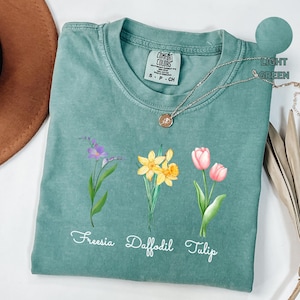 May include: A light green t-shirt with a faded look features three floral illustrations: a freesia, a daffodil, and a tulip. The shirt has the words "Freesia Daffodil Tulip" printed below the flowers. The shirt also has a small tag with the words "Comfort Colors" printed on it.