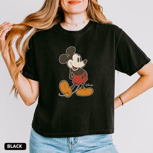 Puede incluir: Camiseta negra con un gr&aacute;fico de Mickey Mouse. El personaje de dibujos animados se muestra en su pose cl&aacute;sica, con pantalones cortos rojos, zapatos amarillos y guantes blancos. El logotipo de Disney es visible. La palabra "BLACK" est&aacute; en la parte inferior.