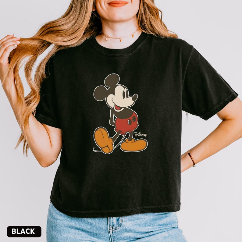 Puede incluir: Camiseta negra con un gr&aacute;fico de Mickey Mouse. El personaje de dibujos animados se muestra en su pose cl&aacute;sica, con pantalones cortos rojos, zapatos amarillos y guantes blancos. El logotipo de Disney es visible. La palabra "BLACK" est&aacute; en la parte inferior.
