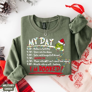 Może przedstawiać: Zielona wojskowa bluza z napisem "MY DAY" i grafiką Grincha w czapce Świętego Mikołaja. Harmonogram obejmuje zajęcia takie jak "Wallow in Self Pity" i "I'M BOOKED!" na czerwono. Widoczny jest srebrny naszyjnik.