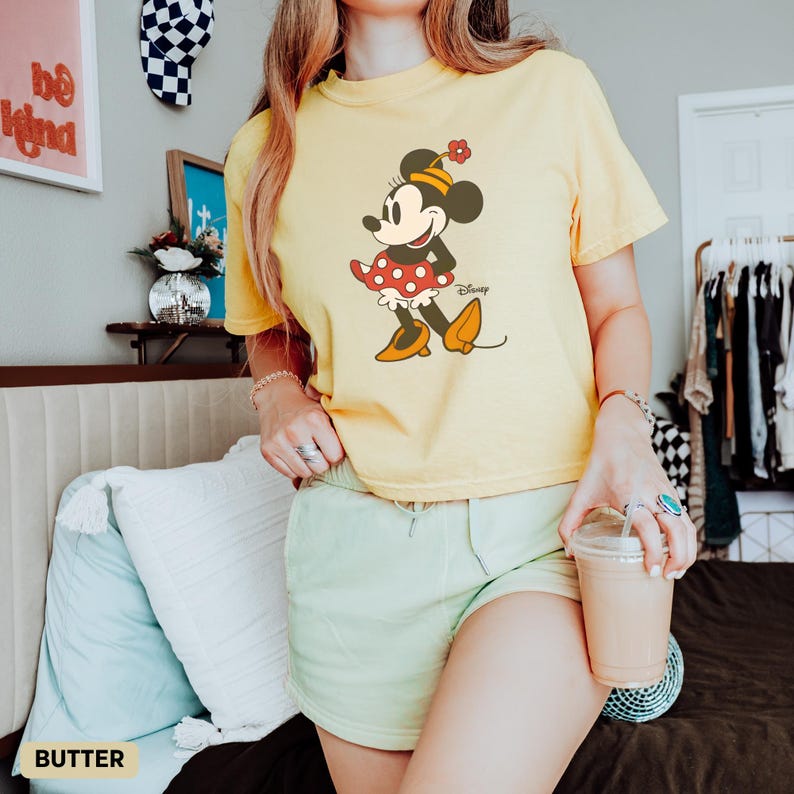 Puede incluir: Una camiseta amarilla con un dise&ntilde;o de Minnie Mouse. El personaje de dibujos animados lleva un vestido de lunares rojos y blancos, zapatos amarillos y una flor en la cabeza. La palabra "Disney" est&aacute; impresa debajo.