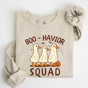 May include: Beige sweatshirt with the text "BOO-HAVIOR SQUAD" in brown. The graphic features three ghost-like ducks with orange beaks and feet, and a small jack-o'-lantern. Bats and stars are also included in the design.