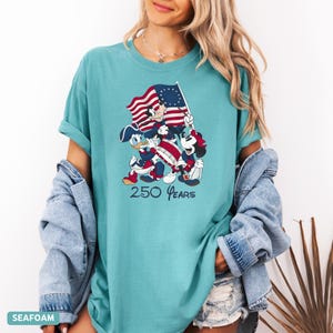 May include: Seafoam green t-shirt featuring a graphic of Mickey Mouse, Donald Duck, and Mickey Mouse playing instruments and holding a flag. The flag has red and white stripes and a blue field with white stars. The text "250 Years" is below the graphic.