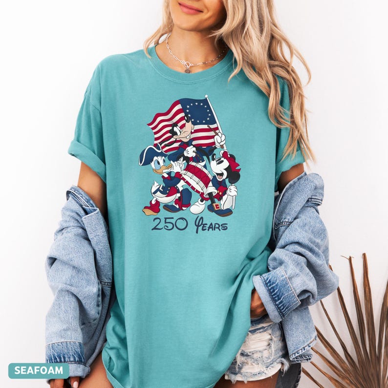 May include: Seafoam green t-shirt featuring a graphic of Mickey Mouse, Donald Duck, and Mickey Mouse playing instruments and holding a flag. The flag has red and white stripes and a blue field with white stars. The text "250 Years" is below the graphic.